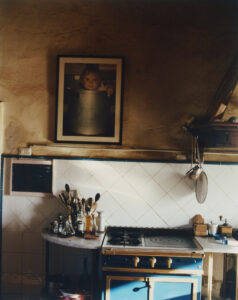 A rustic kitchen with a warm, inviting atmosphere. A blue vintage stove stands against a backdrop of white tiled walls. Utensils and condiments clutter the countertop, adding a lived-in feel. A child's portrait is framed above, adding charm. Light softly filters in, highlighting the textures and creating a cozy, homely vibe with a touch of nostalgia.