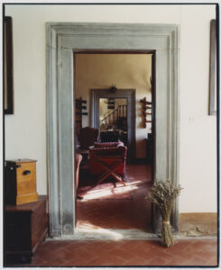 A series of doorways creates an intriguing visual depth, leading into a warmly lit room adorned with leather armchairs and a red ottoman on a patterned rug. Wooden furniture and spiral stairs enhance the cozy ambiance. A bundle of dried lavender leans against one side, adding a rustic touch to the terracotta-tiled floor.