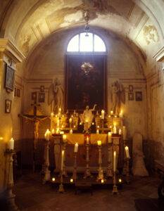 A dimly lit chapel features a majestic altar adorned with numerous glowing candles of varying sizes. Statues of angels flank the scene, while a crucifix and religious imagery decorate the space. Above, the arched ceiling displays faded murals, illuminated by soft light filtering through a window. The atmosphere conveys serenity and reverence in a sacred setting.