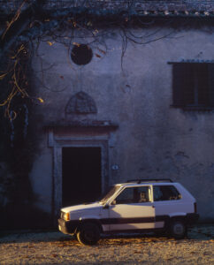A small white car is parked on a gravel path beside an old wall with a weathered door, framed by stone. Above, leafless branches hang with a few yellow leaves catching the light. The building shows signs of age, with a circular vent and closed wooden shutters suggesting history and stillness in the early evening sunlight.
