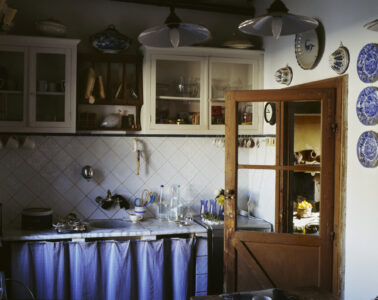 A cozy kitchen features rustic wooden cabinets with glass fronts, displaying dishes and cookware. Decorative plates adorn the walls, and two pendant lights hang from the ceiling. A marble countertop is cluttered with utensils and bottles. The lower cabinetry is hidden behind checkered curtains, and a wooden door is slightly ajar, hinting at another room.