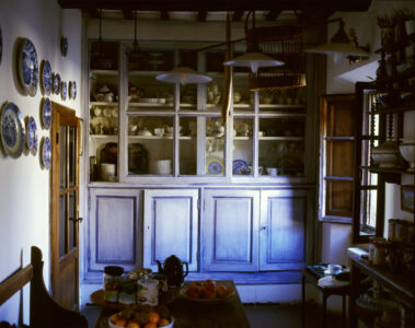 The room has rustic charm, featuring a wooden cabinet with glass doors displaying various dishes and cups. Hanging lights provide a warm glow. Walls are decorated with blue-patterned plates, enhancing the cozy atmosphere. A wooden table holds jars and bowls of fruit, while a wooden door and open window bring in natural light, highlighting the inviting kitchen setting.