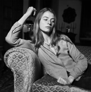 A person with long hair is lounging on a patterned sofa, exhibiting a relaxed posture with an arm resting on the back. The individual wears a buttoned shirt and appears contemplative or introspective. The background is softly lit, featuring furniture and decor that suggest a cozy setting. The overall mood exudes tranquility and reflection.