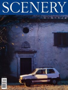 A small car is parked in front of an old stone building with a large wooden door. The building has a round window and closed shutters. Bare branches from a tree, with a few remaining leaves, extend across the scene. Warm sunlight casts long shadows on the ground, creating a tranquil atmosphere.