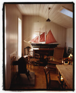 A cozy room features a model ship with red sails atop a cabinet. The wooden floor complements the warm ambiance, with several wooden chairs and a wicker chair arranged around. A dark stove sits near the wall. The ceiling is white with a slanted skylight, allowing sunlight to brighten the space, creating a serene and inviting atmosphere.