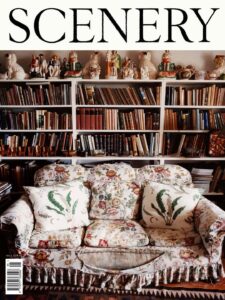 A cosy sitting room is anchored by a floral upholstered sofa layered with botanical cushions. Behind it, overflowing bookshelves stretch across the wall, packed with worn volumes and collected objects. Ceramic figurines line the upper shelves, adding character and nostalgia. Warm tones, dense textures, and abundant details create an inviting atmosphere that feels lived in, personal, and richly curated.