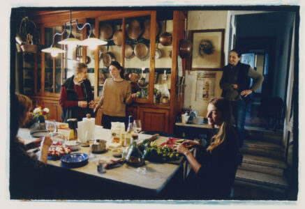 A warm, cozy kitchen setting features a group of five people gathered around a table filled with plates, glasses, and various food items. A woman is chopping vegetables, while others engage in conversation and laughter. Copper pots and pans hang on a wooden cabinet in the background, enhancing the homely atmosphere. Soft lighting adds to the intimate ambiance.