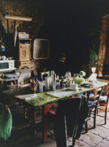 A rustic, dimly lit room features a large, cluttered table surrounded by wooden chairs. The table is covered with a green tablecloth, laden with bottles, containers, and vegetables. Nearby, wooden cabinets hold various items, while a microwave sits atop one. Natural stone walls create a warm, rugged backdrop, with hanging bags adding a touch of color.