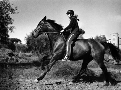A person wearing a helmet rides a horse gracefully through a rustic landscape. The scene captures a sense of freedom and adventure, with trees and vegetation in the background providing a natural setting. The horse displays powerful motion, and the rider maintains a focused posture. A tower partially emerges on the right, adding architectural interest.