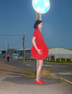 A person stands beside a pole on a road, wearing a bright red outfit that includes a uniquely shaped skirt and shoes. The setting appears to be near an industrial area, with buildings in the background. Overhead, the sky is clear, suggesting it's either dawn or dusk. The scene creates a striking contrast between the attire and the surroundings.