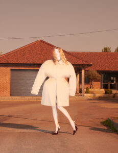 A person stands confidently on a suburban street, wearing an oversized, pale-colored coat. Their high heels accentuate a poised posture. The background features a brick house with a sloped roof and a garage, under a warm, diffuse light that casts long shadows. Sparse greenery punctuates the scene, adding a touch of nature to the residential setting.
