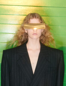A person stands against a vibrant green background, wearing a pinstriped blazer with exaggerated shoulders. The individual sports curly hair and dons a bold, futuristic golden visor that obscures their eyes. The overall aesthetic is striking and avant-garde, blending elements of fashion and science fiction with a minimalist approach.