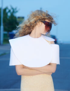 A woman with wavy blonde hair stands with arms crossed, wearing a large pair of dark sunglasses and a distinctive white, sculptural garment resembling a layered collar. Her gaze is directed to the side, and she is placed against an urban backdrop with soft lighting and a blurred background, adding depth to the visual composition.