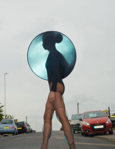 A person walks along a street, wearing a unique outfit with sheer, stylish tights. The attire includes a large, circular accessory that casts an ethereal glow around the head and shoulders. Cars are parked alongside the road, and the cloudy sky adds a dramatic backdrop to the scene, creating an intriguing blend of fashion and urban environment.