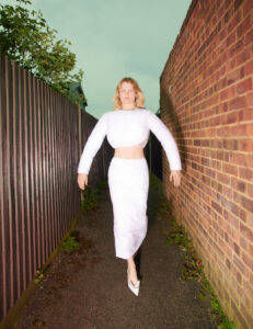 A person poses confidently in a narrow alleyway, flanked by a brick wall and metal fence. Wearing a white, long-sleeved crop top and matching skirt with elegant high heels, the individual exudes a modern, stylish vibe. The setting is enhanced by lush greenery in the background under a light-colored sky, creating a striking contrast with the attire.