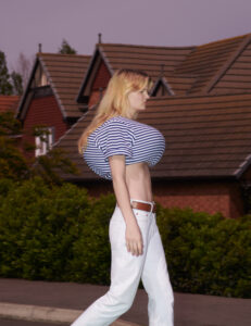 A person with long blonde hair walks in a suburban neighborhood. They wear a blue and white striped crop top and white pants. The background features red-brick houses with tiled roofs and green shrubbery. The scene is set against a clear sky, adding a sense of tranquility. The overall atmosphere is unusual due to exaggerated proportions.