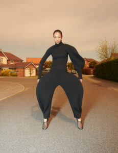 A person stands on a suburban street, wearing an all-black, unusually shaped outfit with large, rounded pant legs and a fitted top. The background features houses, a neatly trimmed hedge, and a muted sky suggesting early morning or evening light. The person's stance is confident and poised, blending fashion-forward aesthetics with a residential setting.