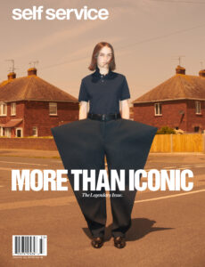 A person stands confidently at a suburban crossroads, wearing a black polo and oversized, dramatically wide black pants that contrast starkly with the setting. Behind them, two brick houses with tiled roofs and chimneys frame the scene. The sky is a muted ochre, enhancing the overall surreal and fashion-forward aesthetic. Shadows stretch across the pavement.