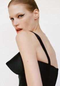 A person with slicked-back hair gazes over their shoulder with a confident expression. They wear bold, dramatic eyeliner that accentuates their eyes. The outfit is elegant, featuring thin straps and a structured design. The overall mood is poised and intense, with a focus on the person's striking makeup and fashion-forward appearance, set against a neutral background.