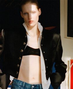 A person stands confidently with a direct gaze, wearing a black jacket with metallic buttons over a white shirt and dark crop top. The top is unbuttoned to reveal a hint of midriff. Blue jeans contribute to a casual yet chic ensemble. Soft lighting highlights their features, creating a contrast with a dark, indistinct background.