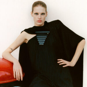 A person with a poised expression leans against a red object, showcasing a sleek, edgy outfit with a black geometric accessory. The ensemble features one sleeveless arm, revealing a tattoo. Hair is styled back, emphasizing bold eye makeup. The background is white and textured, adding contrast and highlighting the striking fashion and commanding presence of the individual.