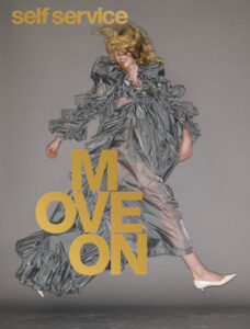 A woman is captured mid-motion, her body lifted as if dancing or leaping. She wears a voluminous, metallic grey dress with exaggerated ruffles and texture that billow around her. Her blonde hair whips across her face, partially obscuring her features. One foot is pointed forward in a low heel, adding elegance to the dynamic, playful energy against a neutral backdrop.