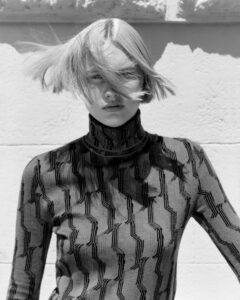 A person with short blonde hair is captured in a candid moment, their hair blowing across their face. They wear a high-neck, patterned sweater, featuring geometric designs with contrasting elements. The play of light and shadows creates an intriguing effect on the garment's texture, enhancing the overall modern and artistic composition against a plain background.