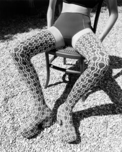 A person is seated on a chair outdoors on a gravel surface. They are wearing high-waisted shorts and bold, patterned thigh-high boots featuring a geometric design. The boots have high heels and extend over the knees. The composition captures a play of light and shadows, highlighting the intricate patterns and textures of the attire.