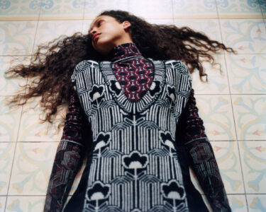 A person with curly hair lies on the floor, gazing sideways. The individual wears a patterned garment with intricate, geometric designs featuring black, white, and deep red hues. The background consists of tiled flooring with soft, muted colors and elaborate designs. The contrast between the attire and tiles creates an artistic, harmonious visual.
