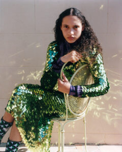 A person with long, curly hair sits on a metal chair. They wear a striking, shimmering dress adorned with large, green sequins that reflect the light, creating a dynamic visual effect. The outfit is paired with black and white socks. The setting appears to be outdoors, with soft natural lighting casting gentle shadows on a neutral background.