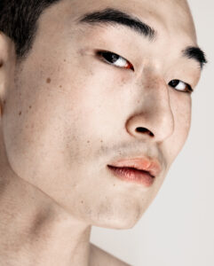 A close-up view features a person's face, highlighting smooth skin with visible freckles and a slightly textured surface. The focus is on one eye, framed by dark eyebrows, and the lips, which have a natural tone. The expression is neutral, with a soft gaze. The composition uses soft lighting to emphasize facial details and contours.