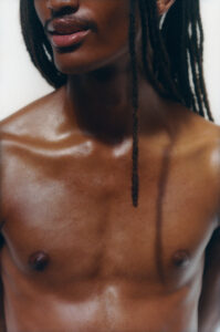 A person with long, dark hair appears from the shoulders upward. Their skin has a smooth, radiant glow, and their expression is relaxed yet confident. A single braid hangs over their left shoulder, highlighting their prominent collarbone. The lighting gently accentuates the contours of their upper chest area, focusing on the person's natural beauty and strength.