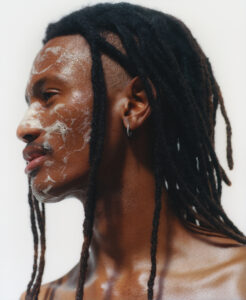 A person with long, dark dreadlocks is facing sideways. The skin tone appears warm and smooth, highlighted by soft lighting. White, creamy texture is applied on the face, adding an artistic element. A small hoop earring is visible on the left ear. The overall mood suggests a blend of creativity and natural beauty.