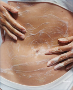 A close-up view showcases two hands gently applying a creamy, white lotion on a smooth midriff. The texture appears rich and moisturizing, creating subtle, swirling patterns on the skin. The lighting highlights the natural skin tone and creamy product, suggesting a skincare or body treatment focused on hydration and care. The overall composition conveys a sense of wellness and body positivity.