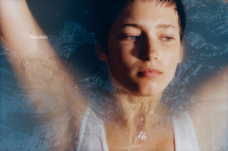 A person with short hair floats in water, arms outstretched. The face is relaxed and gazes upward, capturing a serene and contemplative moment. Light reflects gently on the water’s surface, creating a peaceful ambiance. The person's white top contrasts softly against the blue hue of the water, emphasizing the tranquility of the scene.