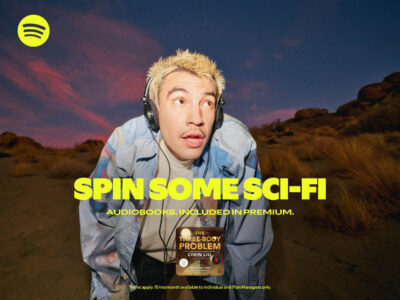 A person with bleached hair and a mustache wears headphones, peering into the distance. Their expression is one of intrigue or surprise. Behind them, a dusk sky with wispy clouds and rocky terrain creates a dramatic backdrop. They sport a pastel-colored jacket over a t-shirt, standing amidst a desert-like setting with sparse vegetation.