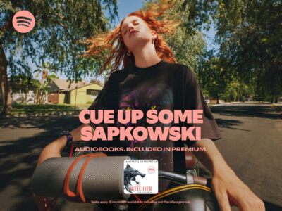 A person with flowing red hair closes their eyes while riding a bicycle on a sunny, tree-lined street. They wear a dark shirt featuring a colorful design. A cylindrical speaker is strapped to the handlebars. The background includes houses and lush green foliage, creating a serene suburban setting. Shadows cast by the trees add contrast to the sunny day.