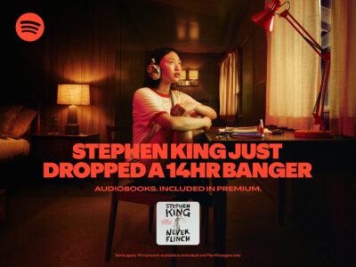 A person wearing headphones sits at a desk, surrounded by warm, ambient lighting. The room features a lamp with a red shade, curtains, and a neatly organized tabletop with stationery items. In the background, a bed with pillows is visible. The overall atmosphere suggests relaxation and focus, enhanced by the cozy and intimate setting.