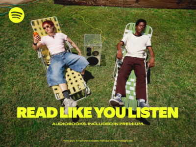 Two people relax on a lush green lawn, each on a sun lounger. One person wears a light pink shirt, denim shorts, and shades, engrossed in their phone. The other is dressed in a white shirt and brown pants, peacefully resting with eyes closed. A boom box sits between them, and a garden hose winds across the grass.