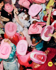 A lively crowd is adorned in colorful attire, featuring pink cowboy hats and various vibrant costumes. Some wear fluffy or sequin-laden outfits, while others accessorize with gloves and glitter. The atmosphere is festive and energetic, as people raise their hands and dance in close proximity, creating a scene filled with dynamic movement and exuberant celebration.