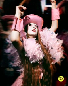 A figure exudes confidence in a flamboyant pink ensemble. The wide-brimmed hat and ruffled, translucent fabric create a theatrical, dreamy aesthetic. Long, flowing hair and heavily styled makeup enhance the dramatic appearance. The dynamic pose captures a sense of motion and flair, while the vibrant colors and playful textures add a surreal, festive atmosphere to the scene.