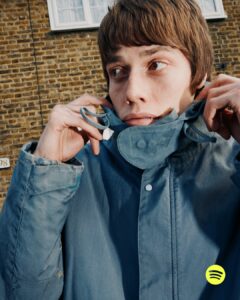 A person with short brown hair fastens their blue jacket's collar, looking slightly to the side. The background features a textured brick wall and a window with white frames. The person’s expression appears thoughtful or contemplative, and the scene has a casual, urban vibe. The lighting casts subtle shadows enhancing the textures.