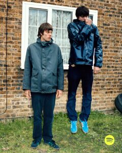 Two individuals stand near a brick wall with a window, dressed in casual, warm clothing. One person appears to be floating, with bright blue shoes, while the other stands on the ground, gazing ahead. The setting is an outdoor area with grass and discarded tires, suggesting an informal, perhaps suburban environment. The scene feels candid and slightly surreal.