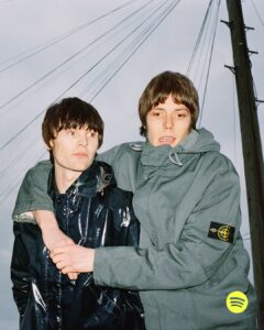 Two individuals, clad in stylish jackets, share a candid moment outdoors. One wears a shiny, dark coat, while the other sports a light gray jacket with a patch on the sleeve. They stand closely, with one's arm draped over the other's shoulder, under a cloudy sky intersected by power lines and poles in the background.