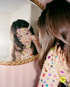 A young woman is gazing into a mirror, her face and upper body adorned with colorful, playful stickers of various shapes like stars, hearts, and butterflies. The scene captures a whimsical and creative expression, with light reflecting off the glossy stickers, creating a vibrant and joyful atmosphere. Her hair cascades naturally, enhancing the imaginative vibe.