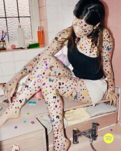 A person sits in a bathtub, covered from head to toe with colorful stickers including stars, butterflies, and other playful shapes. They're wearing a black tank top and white shorts. The surrounding bathroom has pink tiles, a window with textured glass, and various toiletries. The scene is vibrant and whimsical, blending ordinary surroundings with playful creativity.