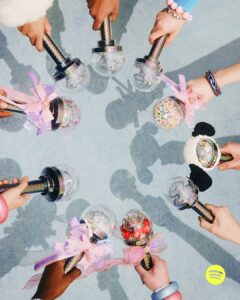 A group of hands holds decorative objects with transparent domes and metallic handles, arranged in a circular formation. Each dome contains various items like cherries and colorful pieces, with some adorned with ribbons and bows. The background is a textured light blue surface, casting soft shadows from the objects, adding depth to the composition.