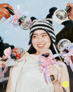 A joyful scene features a smiling individual wearing a black-and-white knitted hat. Surrounding them, several hands hold decorative items with transparent spherical tops, adorned with colorful ribbons. The outdoor background has a clear sky and the edge of a building with greenery. The atmosphere is playful and vibrant, capturing a moment of unity and celebration.