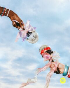 Two hands, each holding a lightstick adorned with decorative bows, reach towards each other against a background of a bright, clear sky. One hand features bracelets, while the other holds a lightstick topped with cherries. The ribbons on the sticks are pastel and shimmery, creating a playful and vibrant atmosphere.