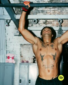 A person performs a pull-up in a gym environment, gripping a bar with both hands. They wear fingerless gloves and have several tattoos on their chest and arms. Their gaze is focused upward, and they are shirtless, wearing dark shorts. Behind them, lockers and a pink stuffed animal stand against a brick wall. Chains hang from the ceiling.